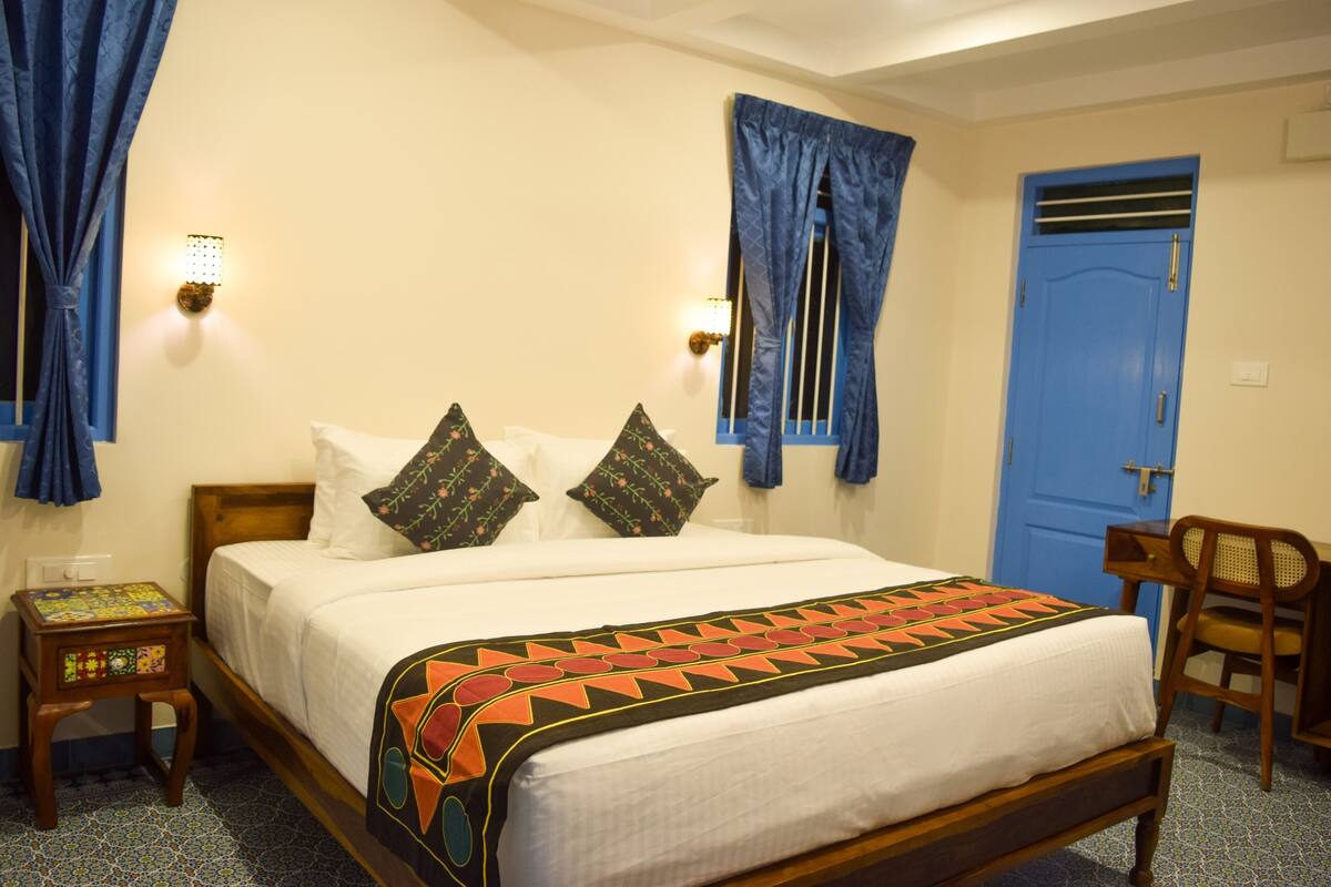 A neatly made bed with a patterned bedspread, two wall-mounted lamps, a blue door, and a small wooden chair.