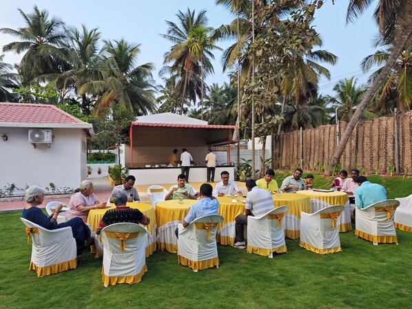 A group of people seated at round tables outdoors with yellow tablecloths and white chairs with yellow sashes, under a covered area with a red roof, surrounded by palm trees. - Ibex Resorts Coimbatore Kakarala (Coimbatore)