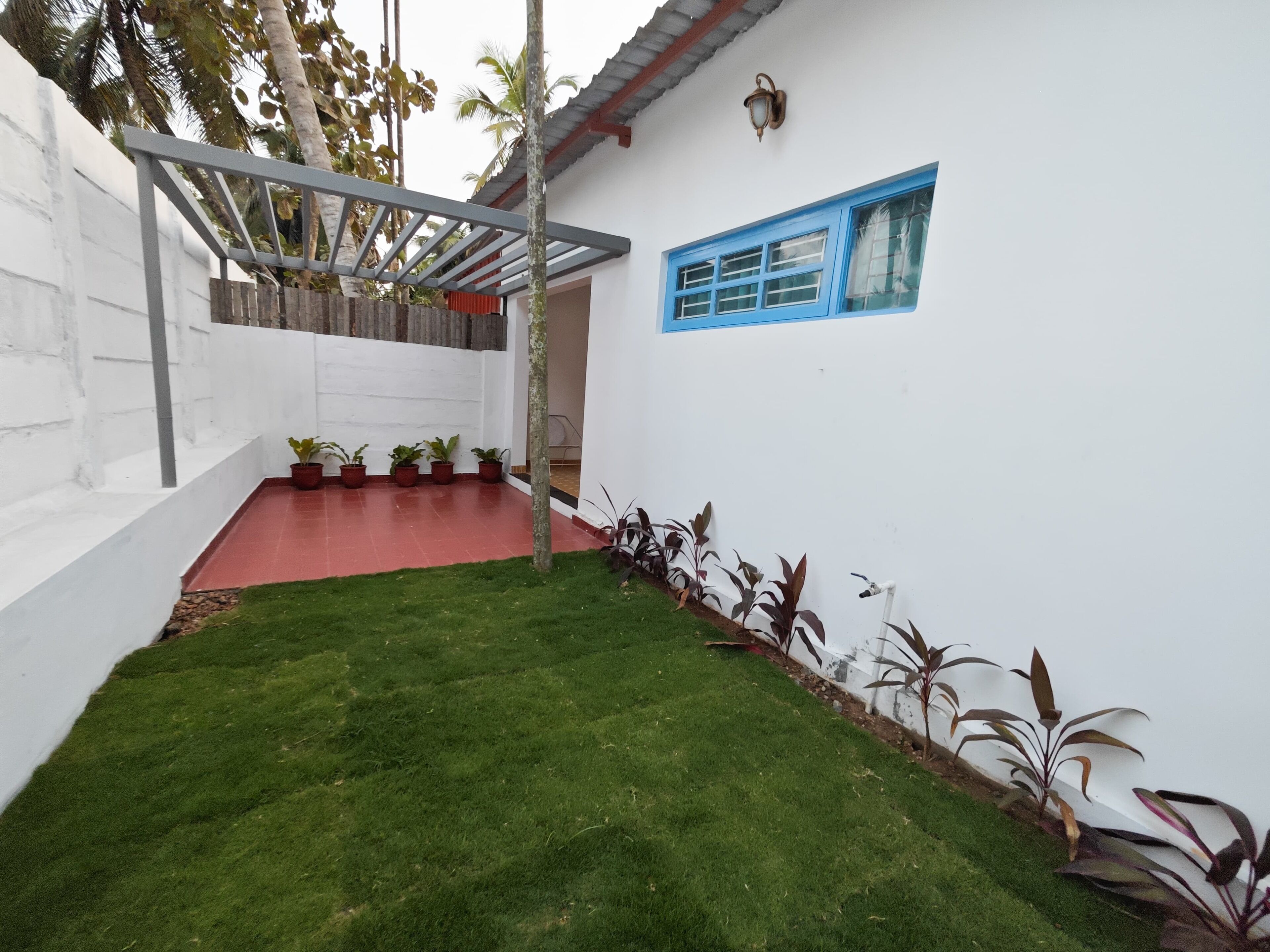 A white building with a red-tiled patio, a blue window, and a pergola with potted plants.