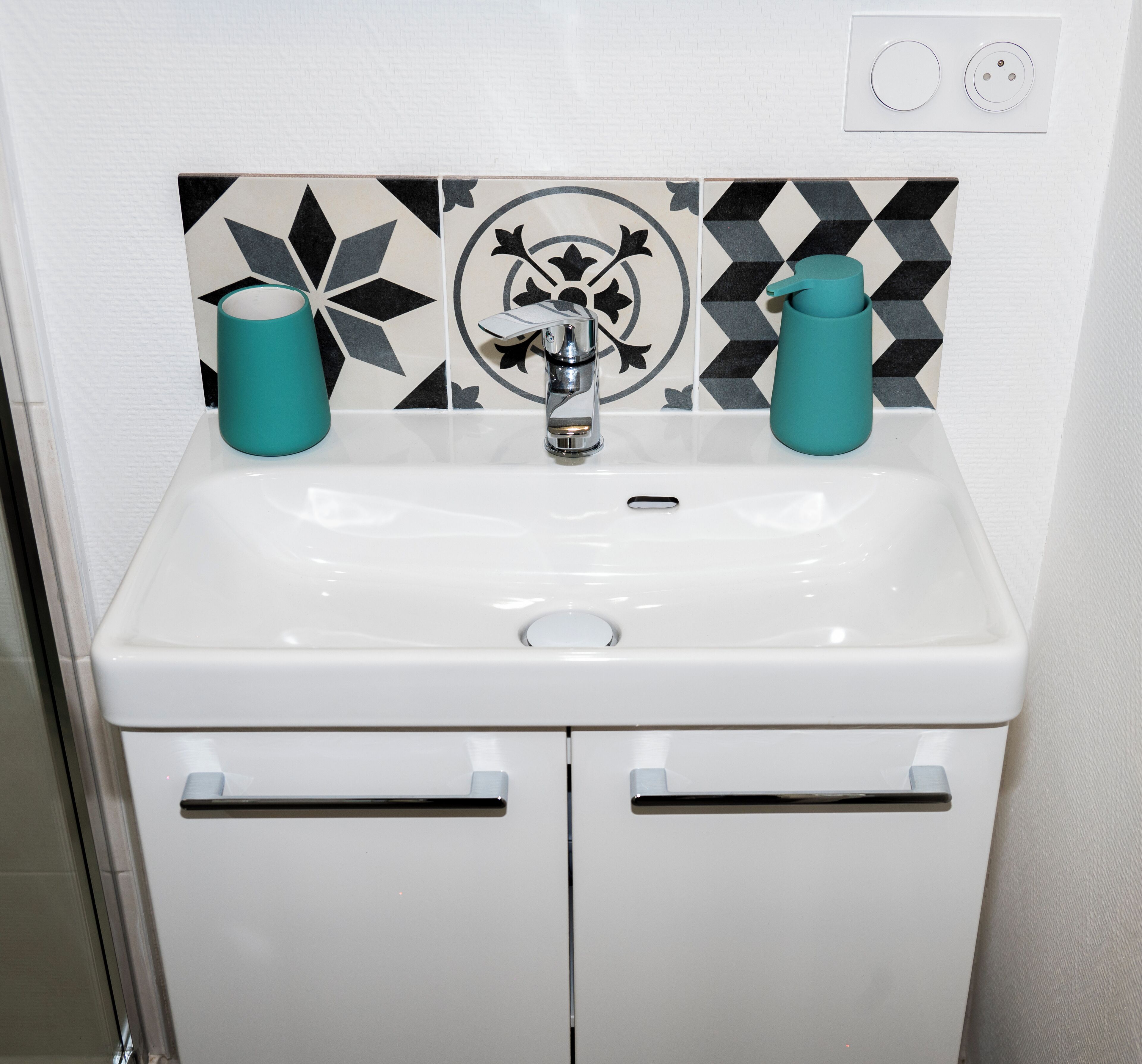 Un lavabo blanc pour la salle de bain, avec deux tasses de couleur turquoise, un robinet moderne et un pare-éclaboussures décoratif en carreaux.