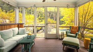 A covered porch with a wooden floor, a green sofa, a wicker table, and a chair with a throw blanket. - Charming VT ski retreat with hot tub, pool, fire pit & endless mountain views (Weston)