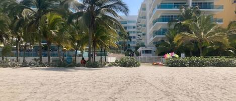 A sandy beach with palm trees and a modern building in the background.