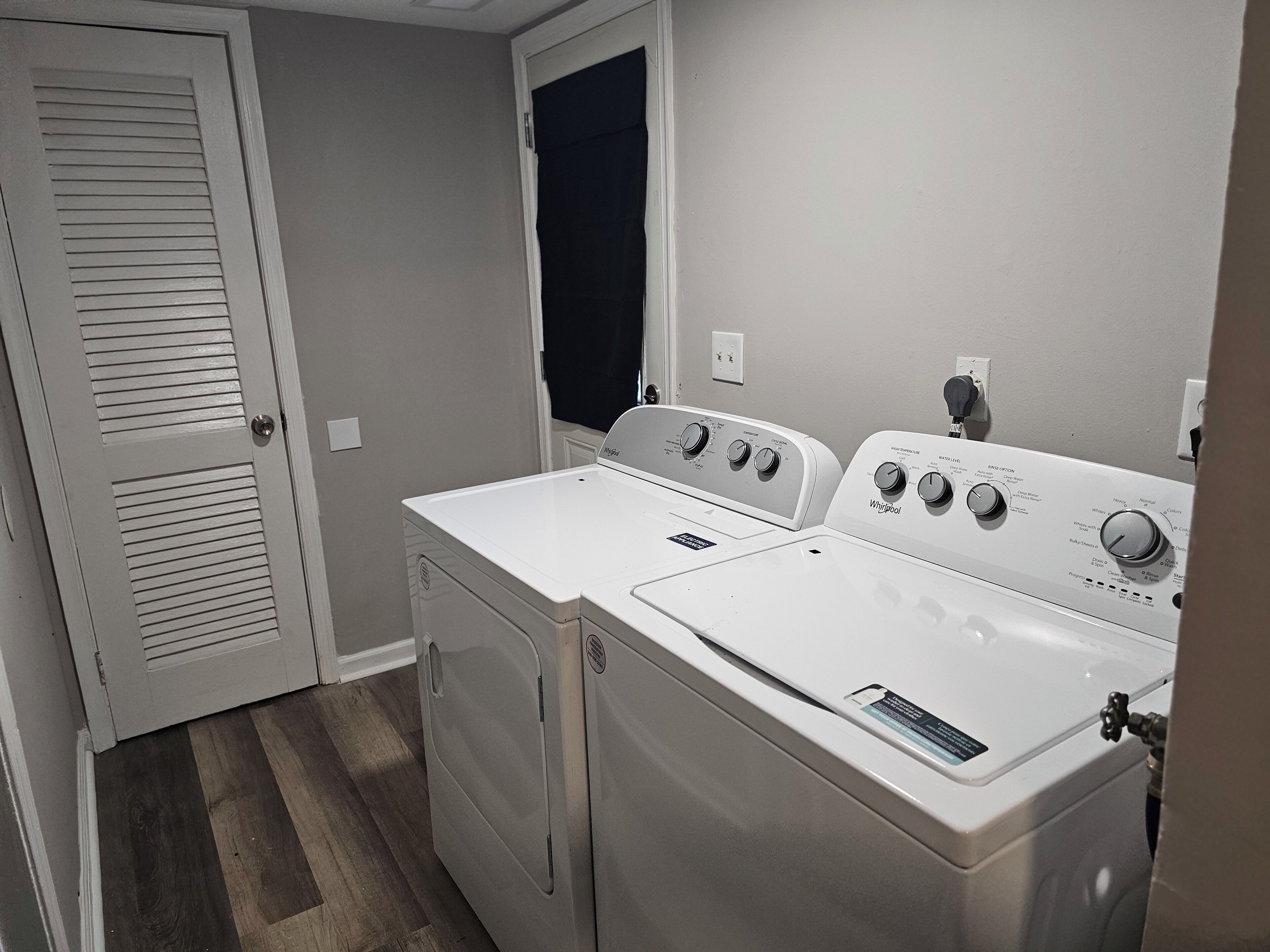 Uma lavanderia com duas máquinas de lavar e secar Whirlpool brancas, uma porta com venezianas e uma cortina preta na janela.