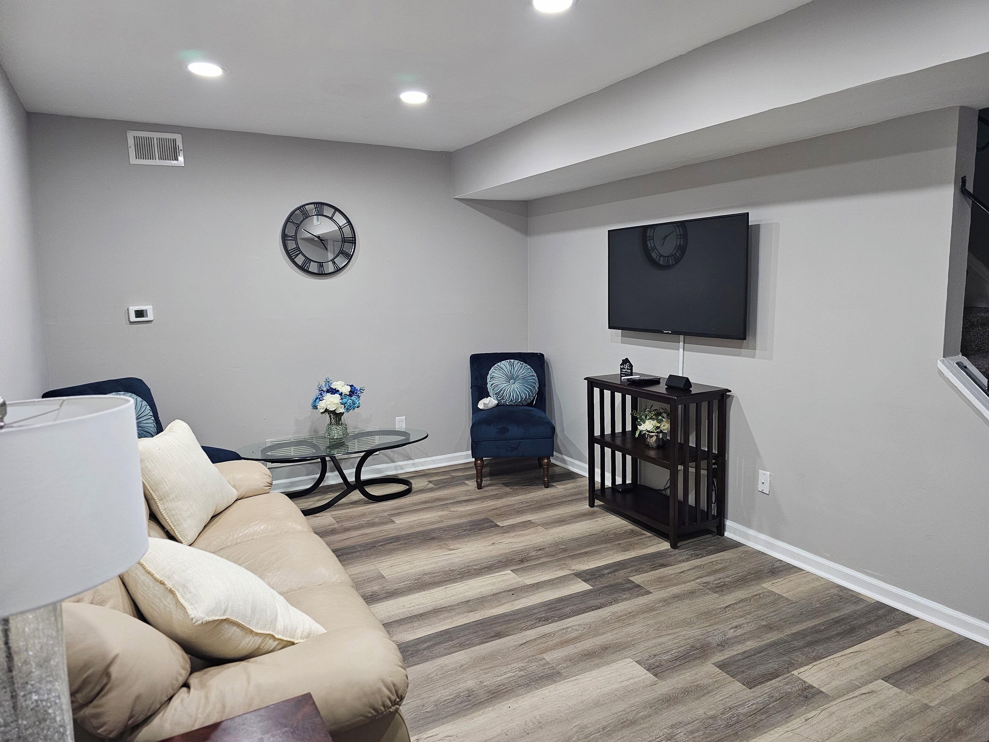 Una sala de estar moderna con un sofá color beige, una mesa de centro de vidrio, un sillón azul y un televisor instalado en la pared.