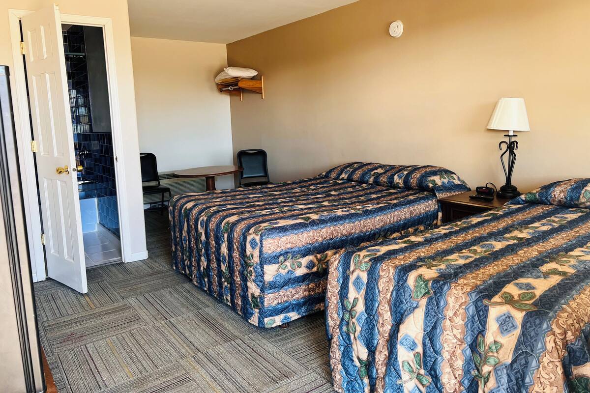 A hotel room with two beds, a desk, a chair, a lamp, and a door leading to a bathroom.
