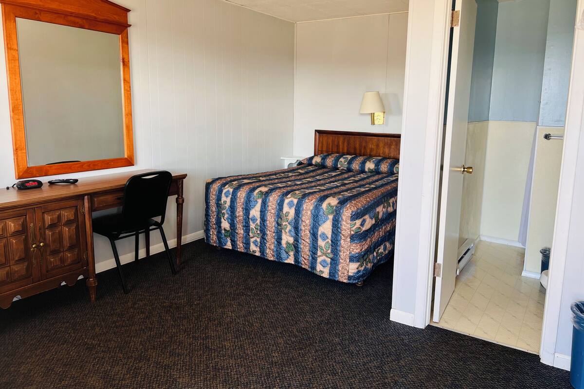 A hotel room with a bed, a desk with a chair, a mirror, and a door leading to a bathroom.