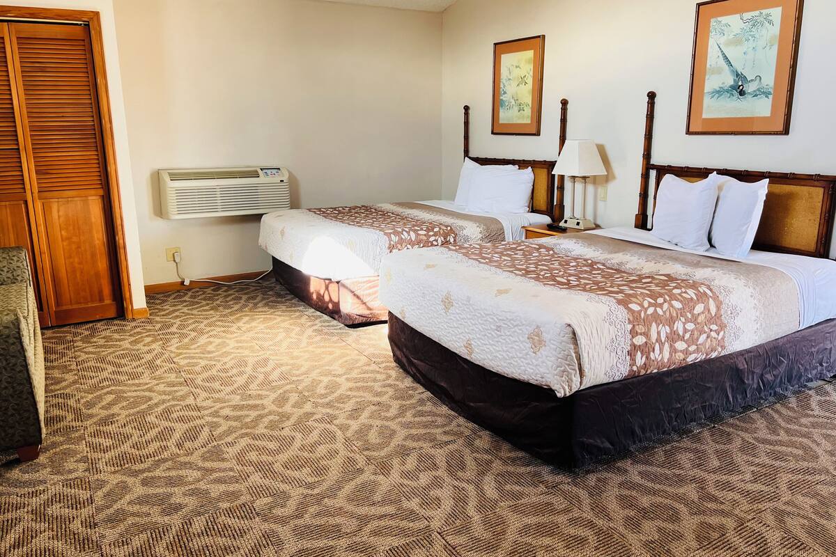 A hotel room with two beds, a wooden door, an air conditioning unit, and two framed artworks on the wall.