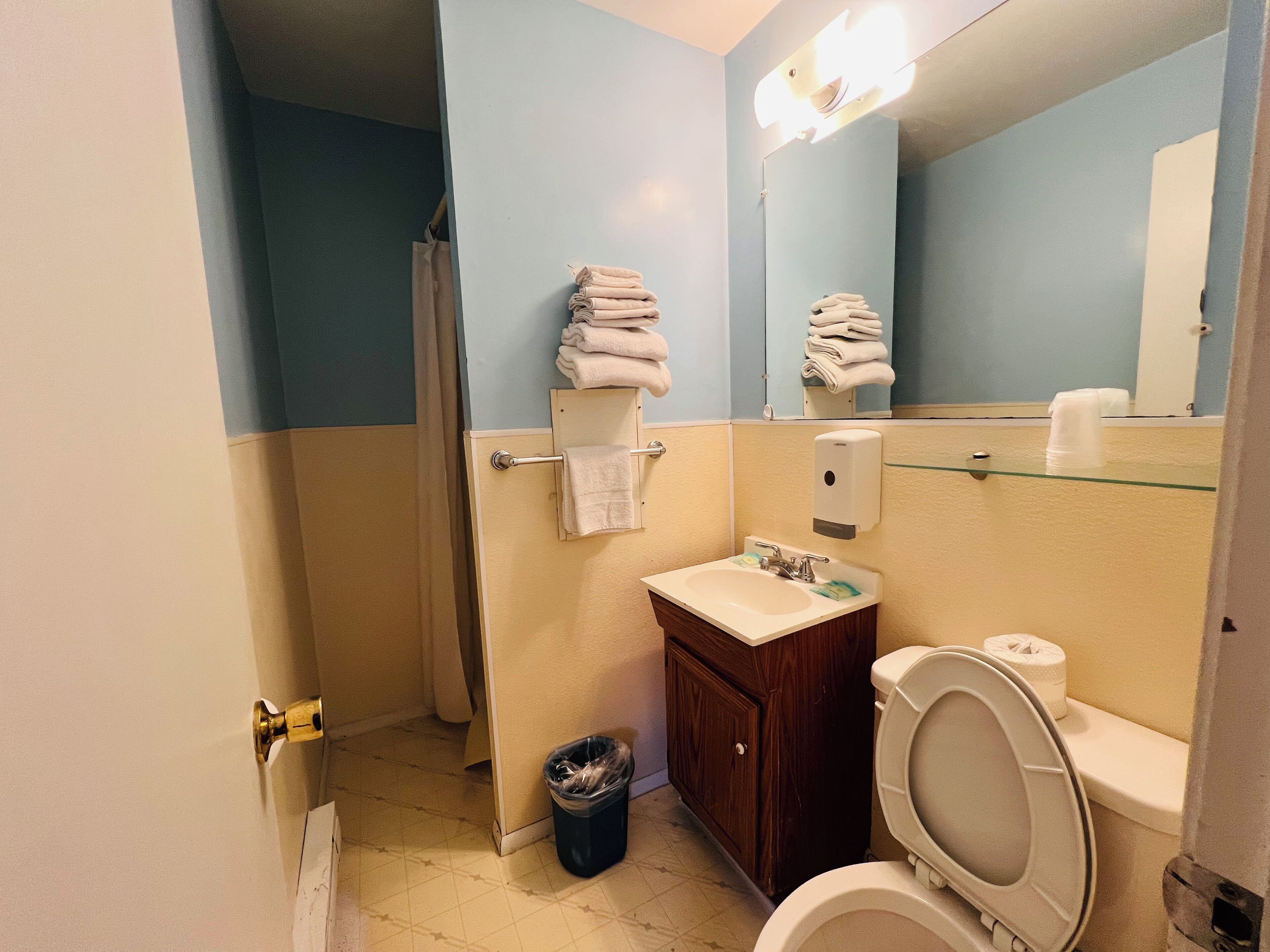 Un baño con lavamanos, inodoro y espejo. Hay toallas colgadas en un perchero y un cubo de basura.
