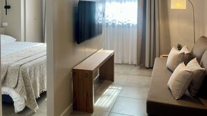 Una habitación de hotel moderna con una cama, un televisor montado en la pared, un banco de madera, un sofá con cojines y un ventanal con cortinas.
