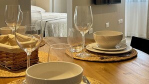 Una mesa de comedor dispuesta con cuencos blancos, copas de vino y cubiertos, con un televisor montado en la pared y una cama a la vista al fondo.