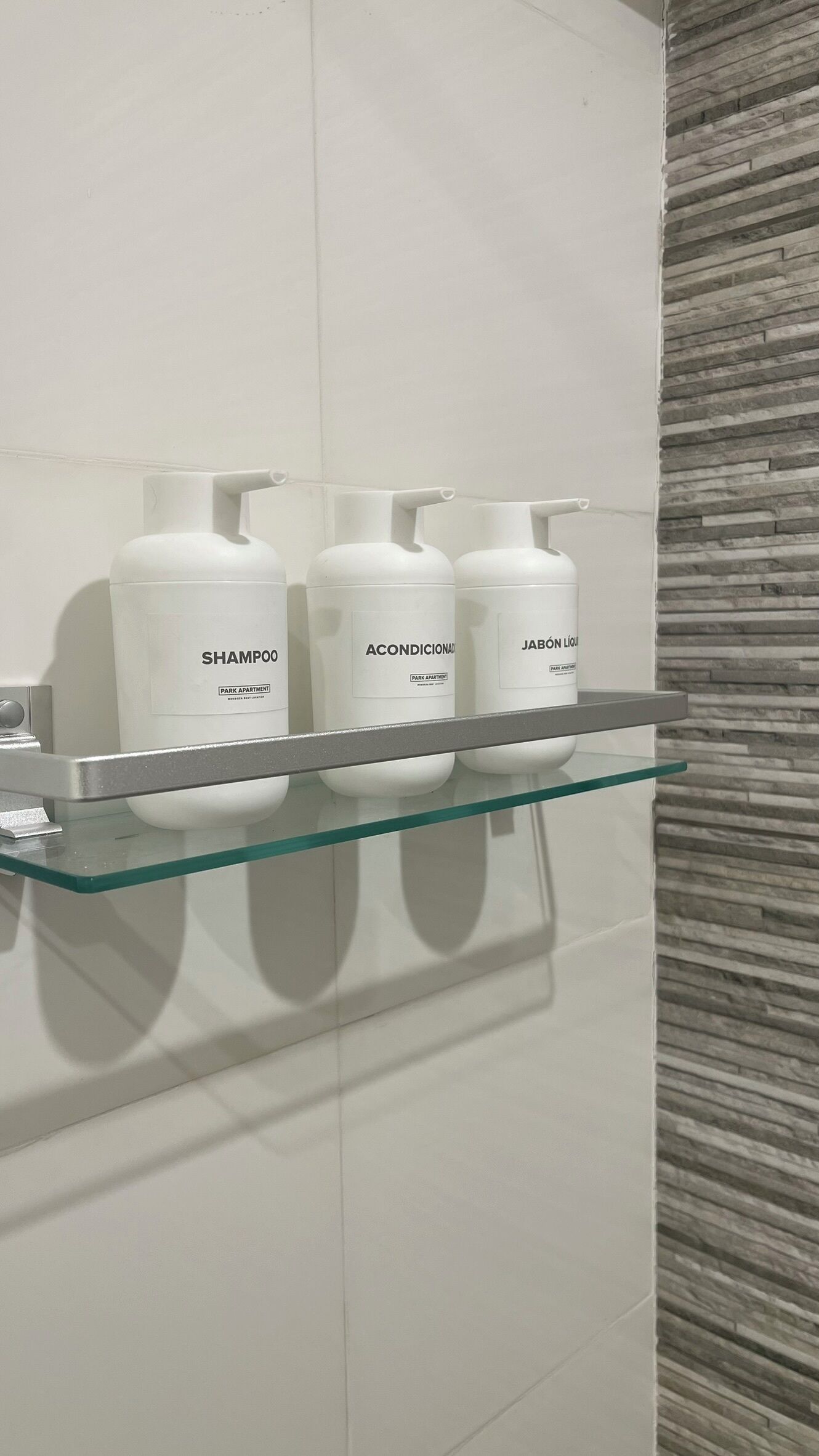 A bathroom with a glass shelf holding three white bottles labeled 'SHAMPOO', 'ACONDICIONADOR', and 'JABÓN LÍQUIDO'.