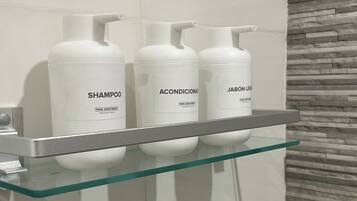 A bathroom with a glass shelf holding three white bottles labeled 'SHAMPOO', 'ACONDICIONADOR', and 'JABÓN LÍQUIDO'.