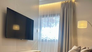 Una sala de estar moderna con un televisor de pantalla plana montado en la pared, un banco de madera, un sofá beige con cojines y un ventanal con cortinas translúcidas.