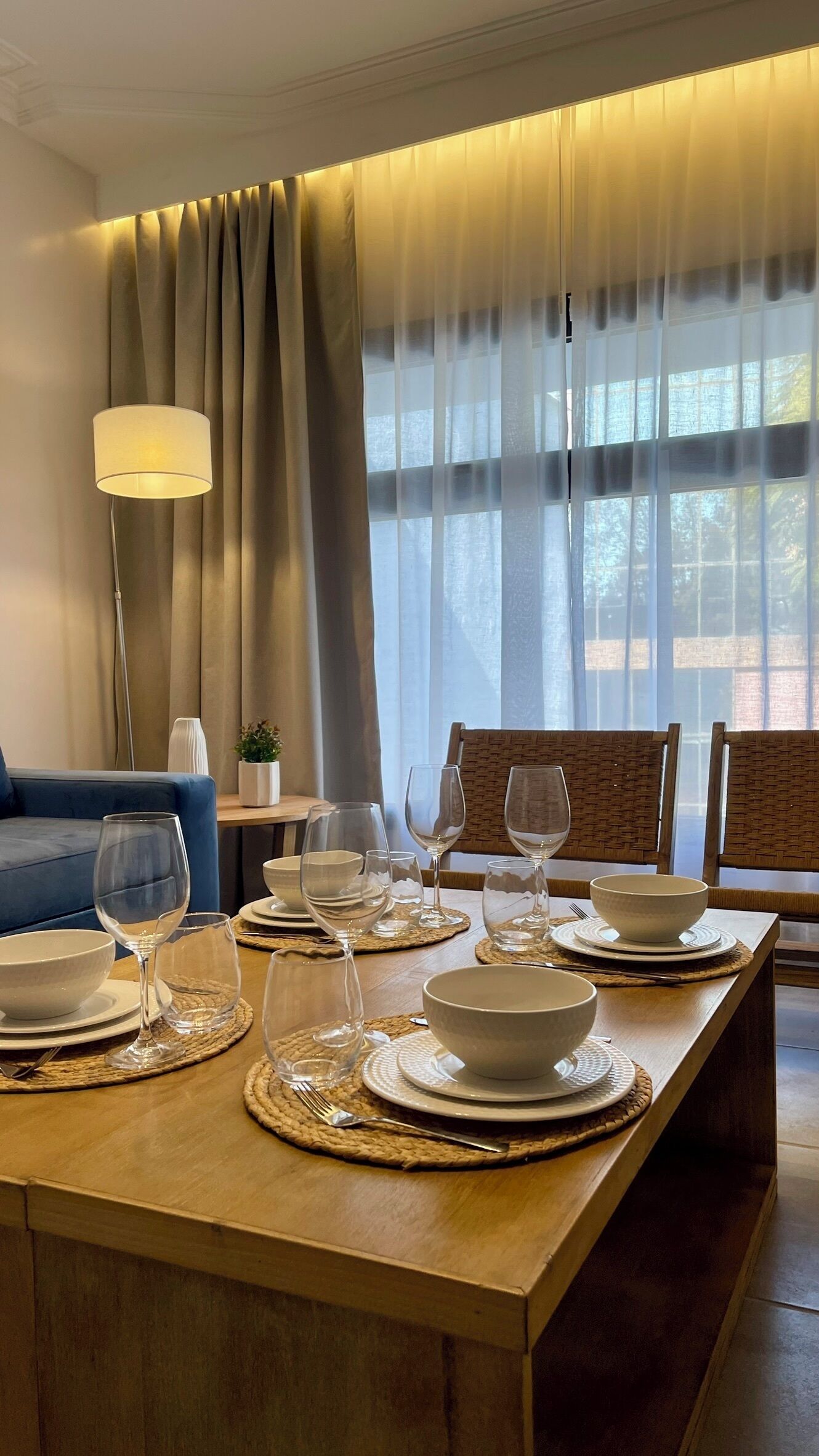 A dining area with a wooden table set for four, including plates, bowls, and wine glasses. There is a blue sofa and a lamp in the background.
