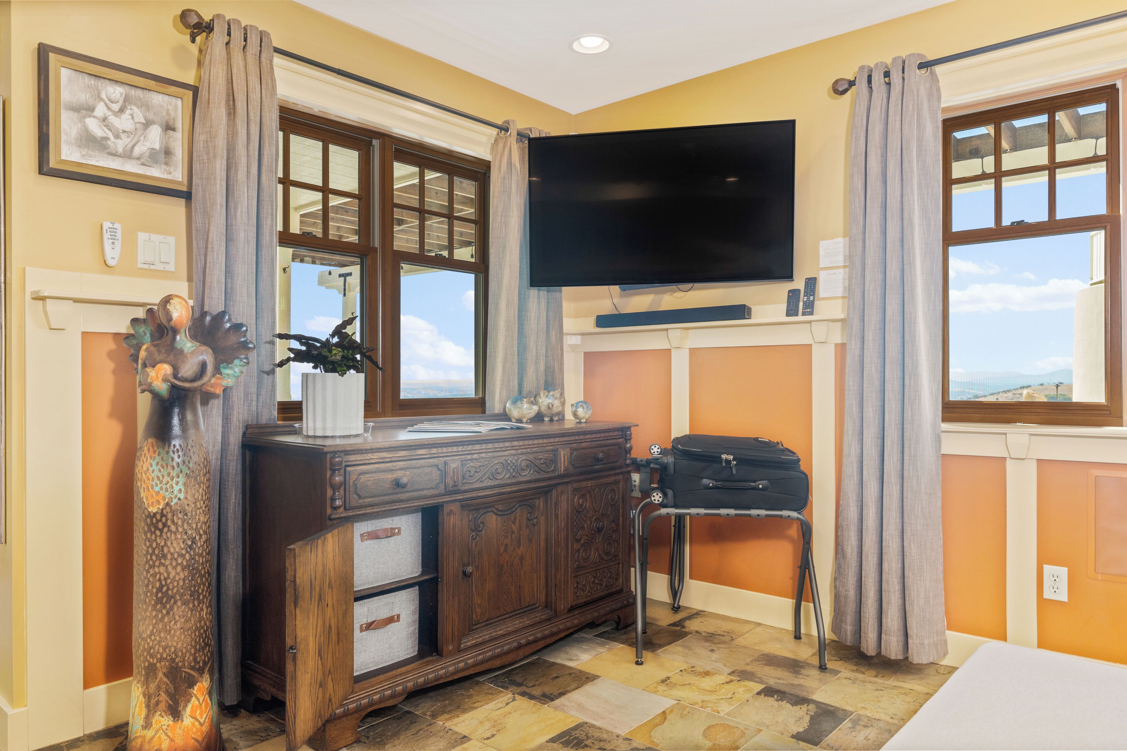 A hotel room with a large flat-screen TV mounted above a wooden dresser, a window with curtains, and a view of the outdoors.