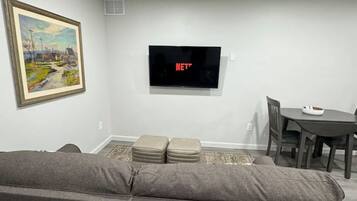A living room with a gray sofa, a painting on the wall, a flat-screen TV, and a dining area with a table and chairs.