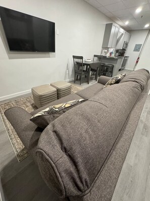 Ein modernes Wohnzimmer mit einer langen, grauen Couch, einem flachbildfernseher an der Wand und einer kleinen Küchenzeile im Hintergrund.