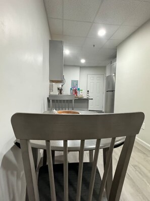 A dining area with a round table and a wooden chair. A kitchen with a refrigerator and cabinets is visible in the background.