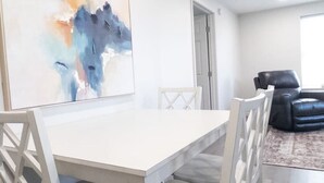 Ett modernt matområde med ett vitt bord och stolar, en stor abstrakt målning på väggen och en svart lädersoffa i bakgrunden.