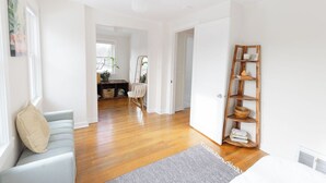 Un espacio de sala luminoso y amplio, con piso de madera, un sofá gris, una estantería de madera y una puerta que da a otra habitación.