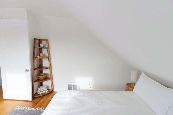 Un dormitorio con techo inclinado, una cama con ropa blanca, una estantería de madera con libros, una mesita de noche con lámpara y una puerta blanca.