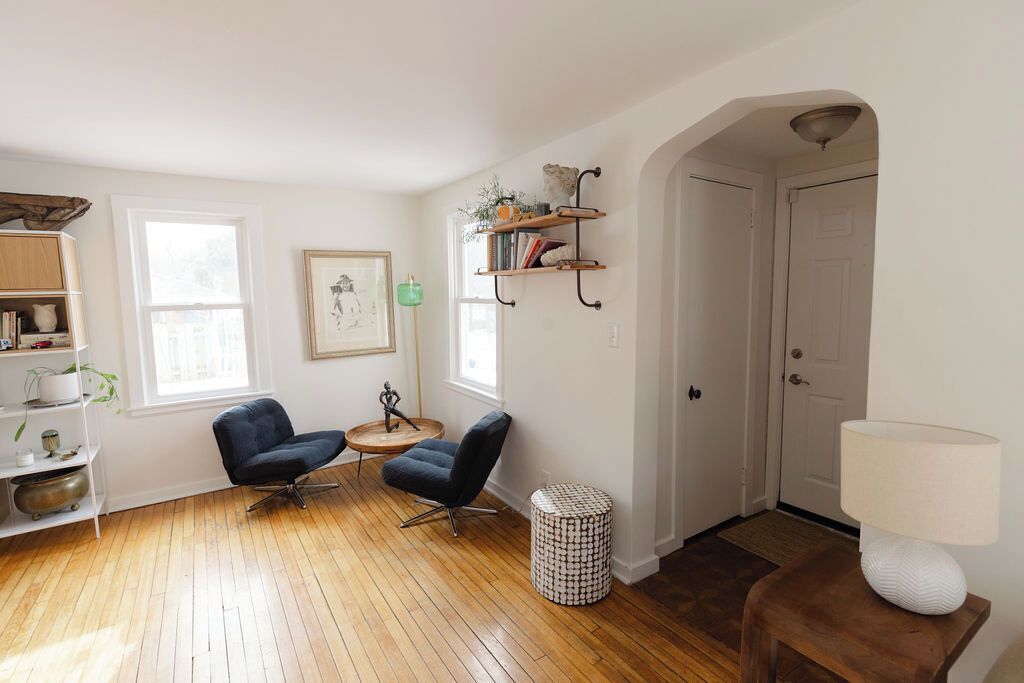 A living room with wooden flooring, two black armchairs, a small round table, a bookshelf, a framed picture, and a door.