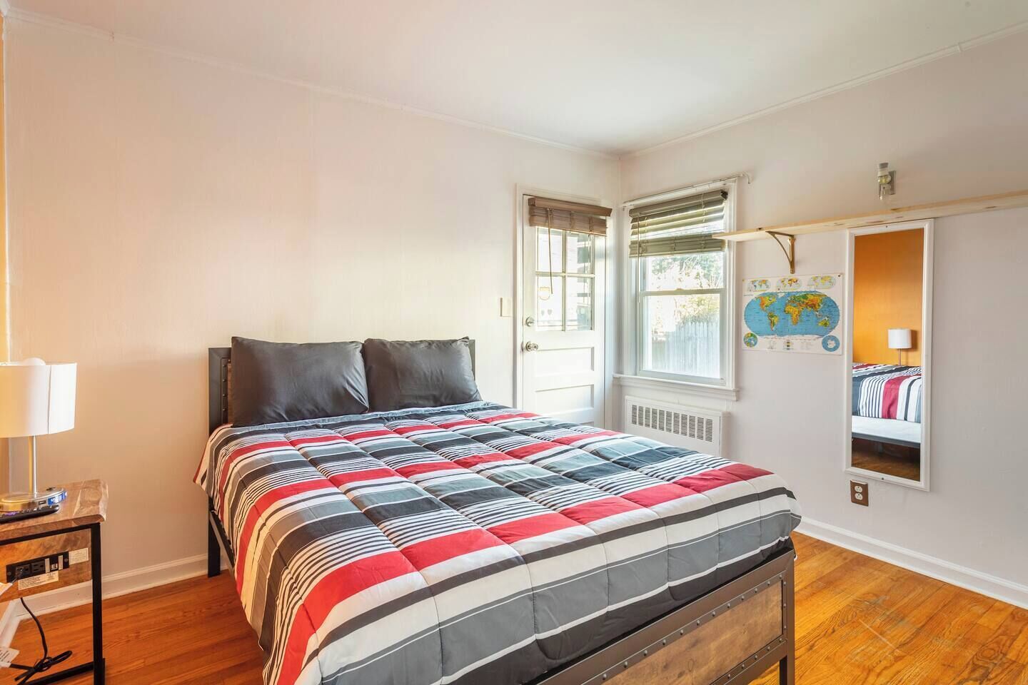 A bedroom with a bed, a nightstand, a window with blinds, a map on the wall, and a mirror.