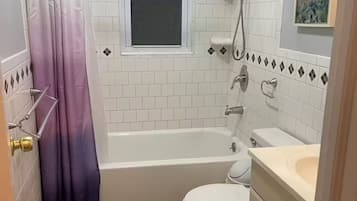 Une salle de bain équipée d’une baignoire, de toilettes et d’un lavabo, avec des carreaux blancs et une fenêtre.