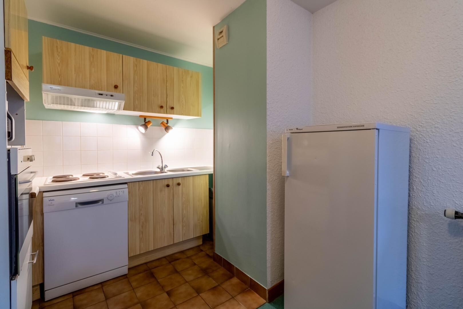 A compact kitchen with wooden cabinets, a white dishwasher, a white refrigerator, and a tiled floor.