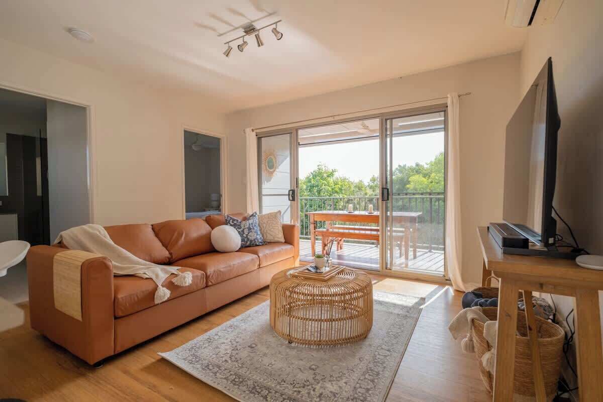 Un salon moderne avec un canapé en cuir marron, un pouf en osier et un téléviseur à écran plat posé sur un meuble en bois.