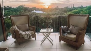 Een balkon met twee rieten fauteuils en een tafeltje, met uitzicht op een weelderig landschap tijdens zonsondergang.