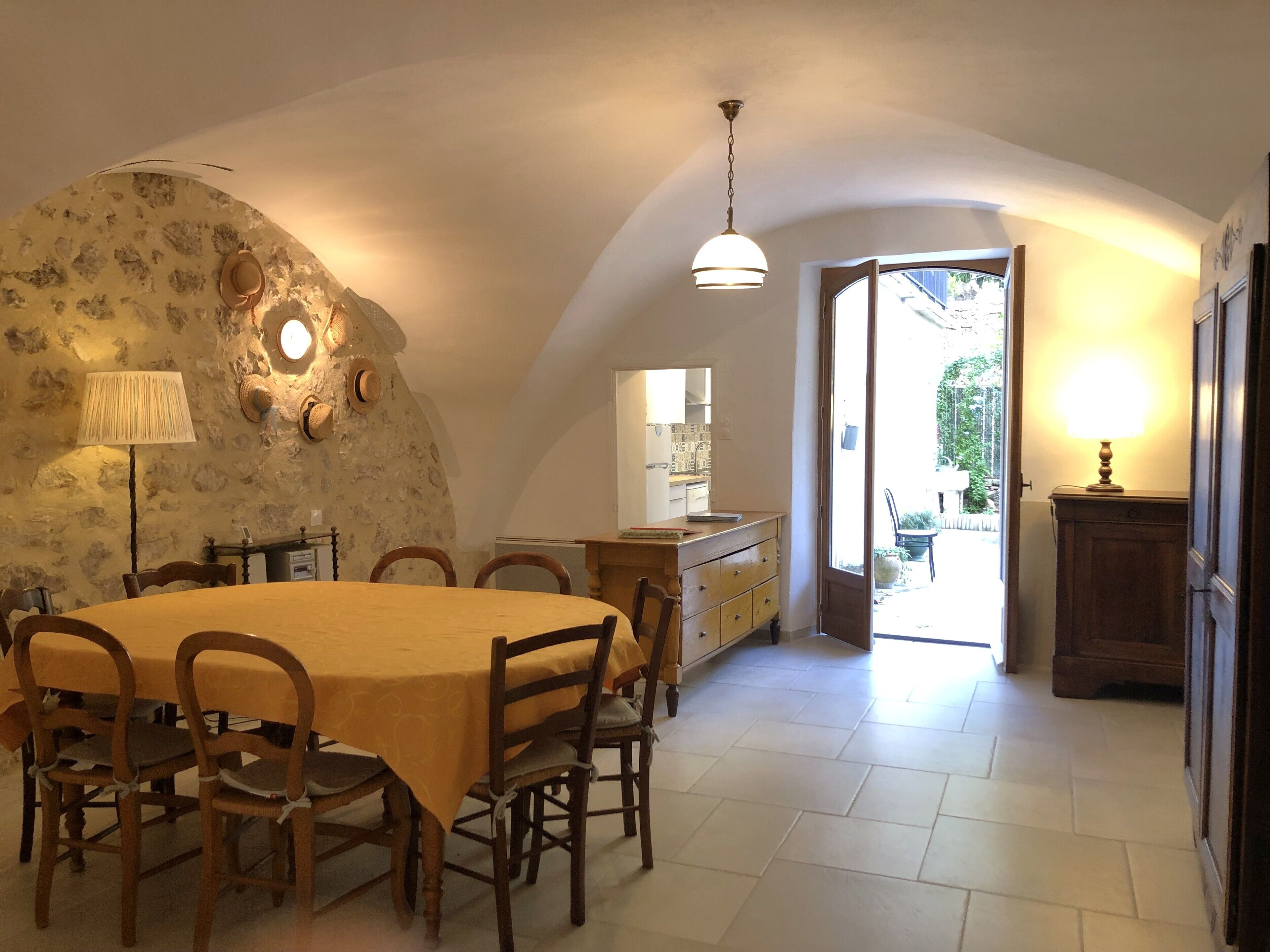 Un coin repas avec une table ronde, des chaises et une lampe. On peut apercevoir la cuisine à travers une porte ouverte.