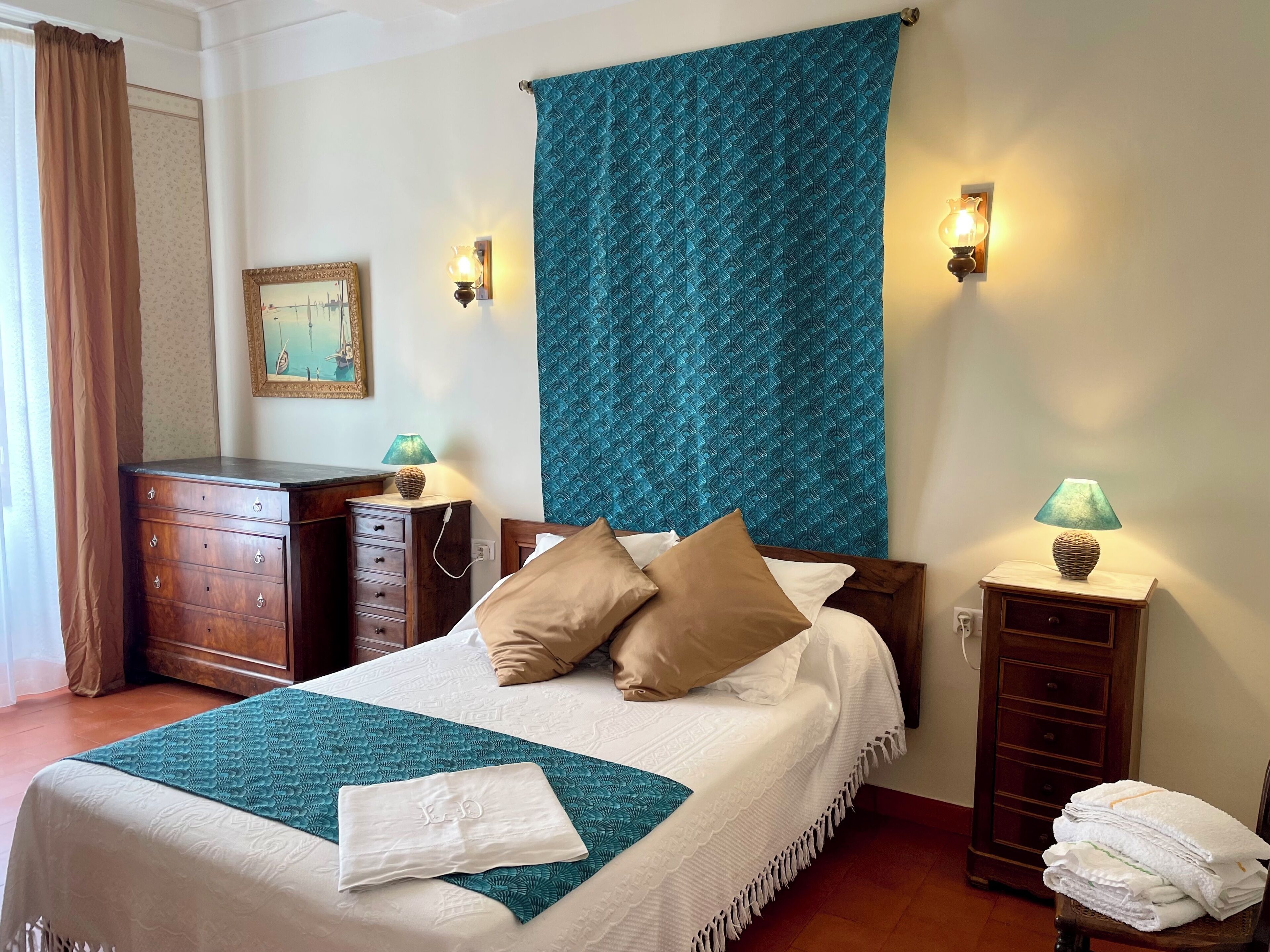 A neatly made bed with a teal and white bedspread, two wall-mounted lamps, a wooden dresser, and a framed picture on the wall.