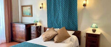 A neatly made bed with a teal and white bedspread, two wall-mounted lamps, a wooden dresser, and a framed picture on the wall.