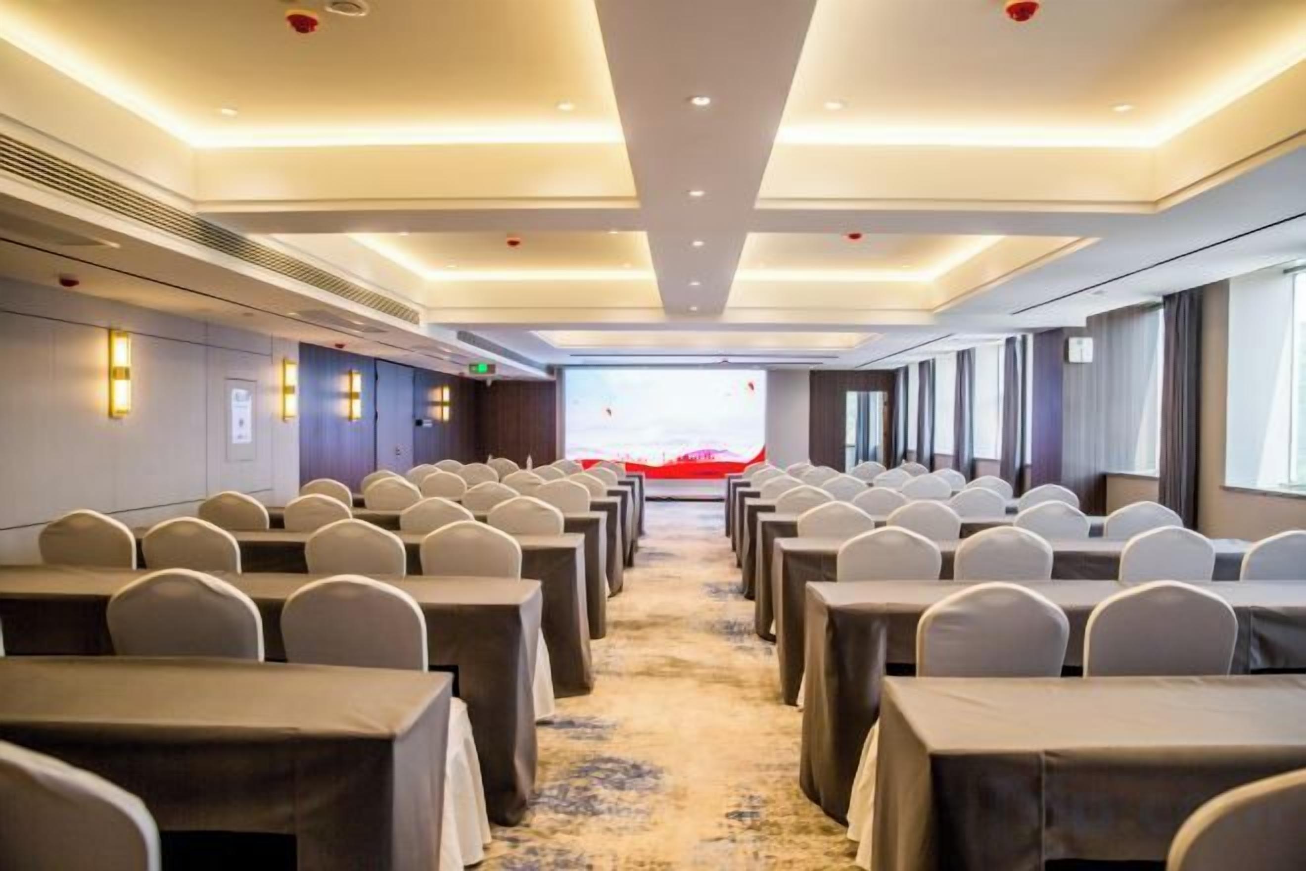 A conference room with rows of tables and chairs, a carpeted floor, and a large screen at the front.