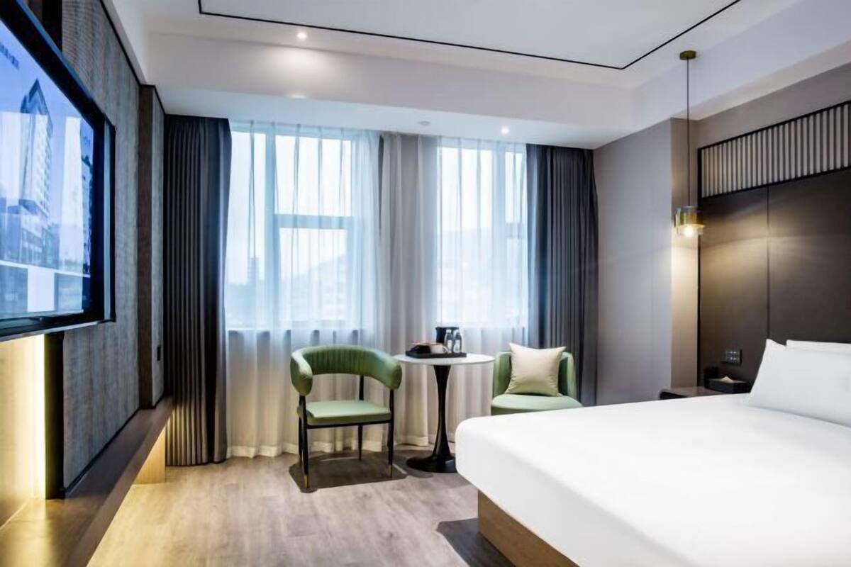 Une chambre d’hôtel avec un grand lit, un téléviseur fixé au mur, une petite table ronde, deux chaises et une vue sur la ville grâce à de grandes fenêtres.