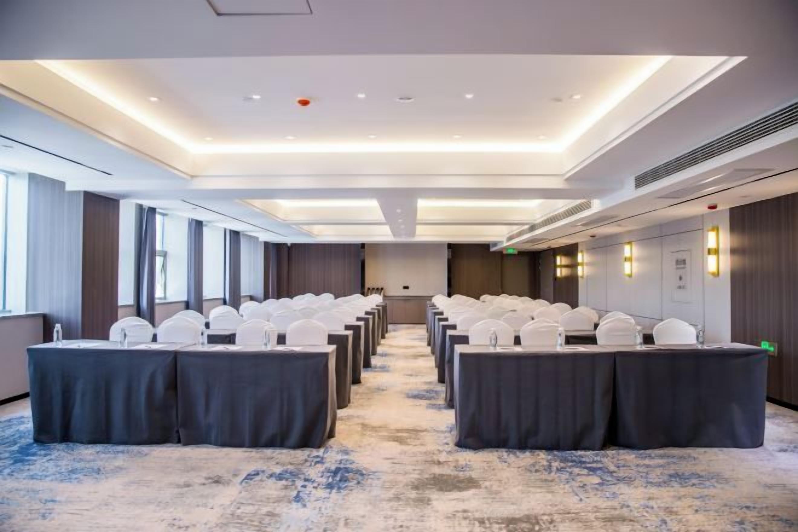A conference room with rows of tables and chairs, a carpeted floor, and a modern ceiling design.