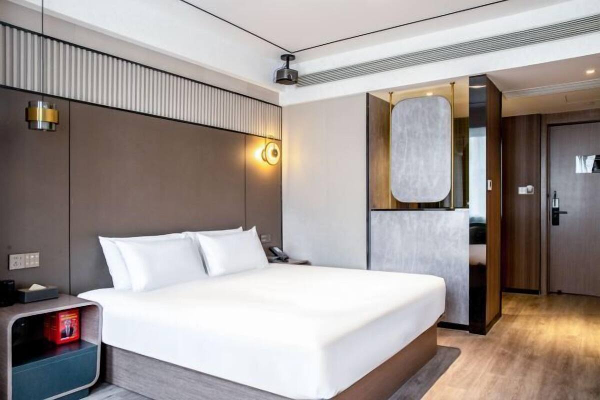 Une chambre d’hôtel moderne avec un grand lit, une table de chevet avec une lampe, un miroir et une porte donnant accès à la salle de bain.