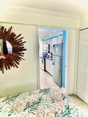 A bedroom with a bed, a decorative mirror, and a view into a kitchen with cabinets and appliances.