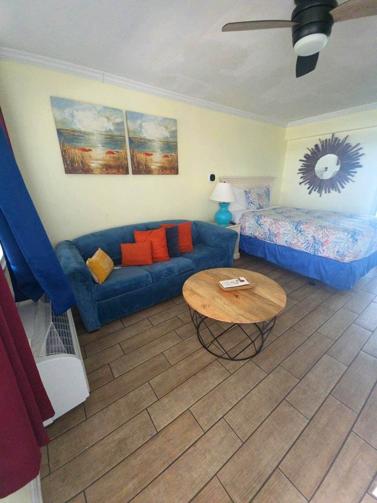A room with a blue sofa, a round wooden table, a bed with a floral bedspread, and two wall-mounted paintings.