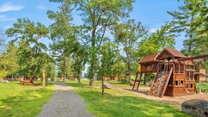 Ein Spielplatz mit einer hölzernen Kletterkonstruktion und einer Rutsche, umgeben von Bäumen und einem Kiesweg.