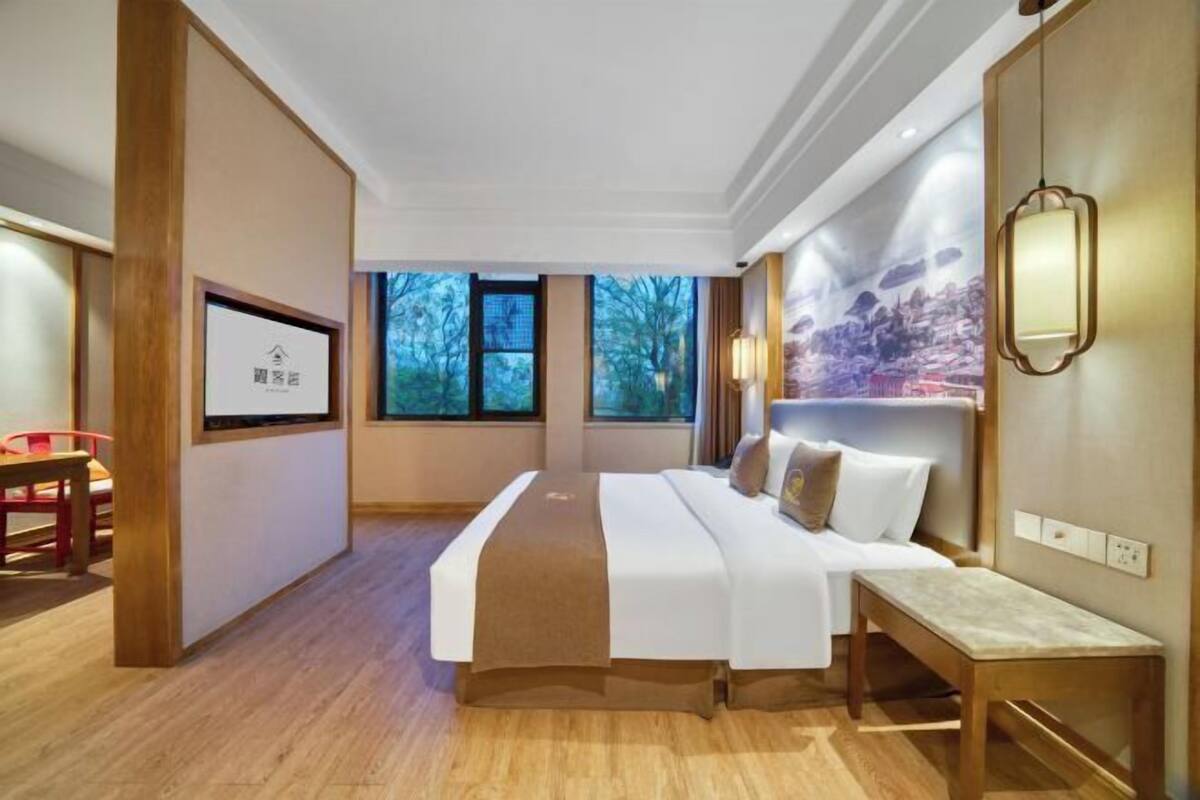 A hotel room with a bed, a desk, and a chair. There are windows with a view of trees.