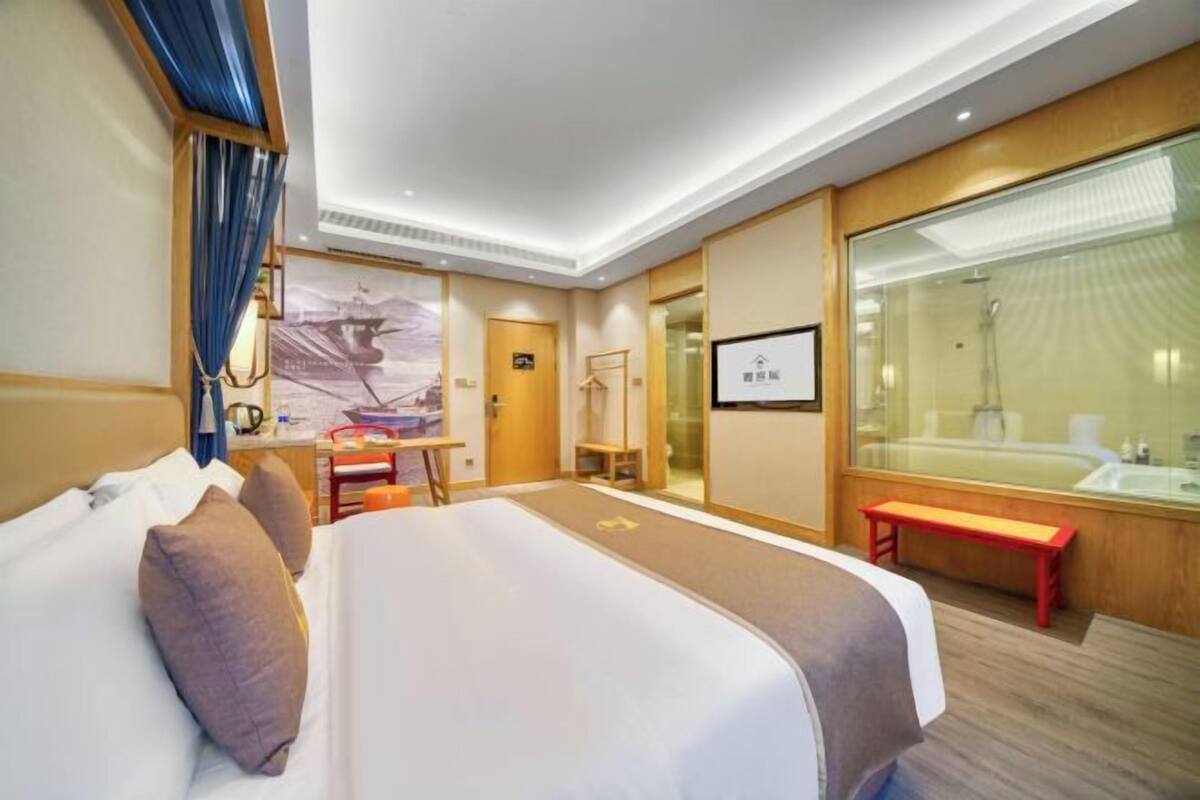 A hotel room with a large bed, a desk, a bench, a shower area, and a large window.