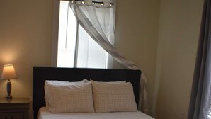 A neatly made bed with a dark headboard, a bedside lamp, and a window with sheer curtains. - Cozy Coastal Getaway (Ingleside)