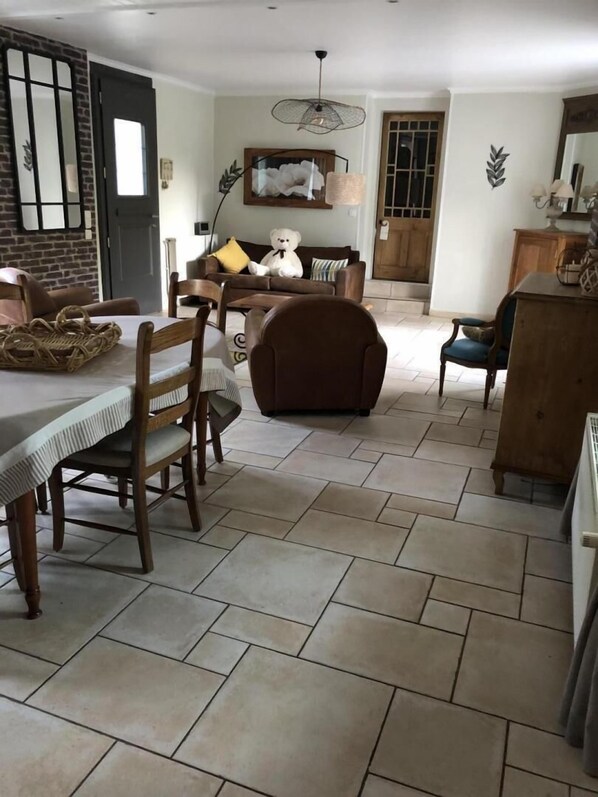 A dining area with a table set for two, a brown sofa, and a teddy bear on the sofa. - Le clos de la Pommeraie (CARLY)