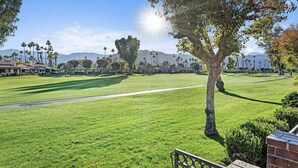 A well-maintained golf course with a clear path, a large tree in the foreground, and a distant view of houses and mountains.