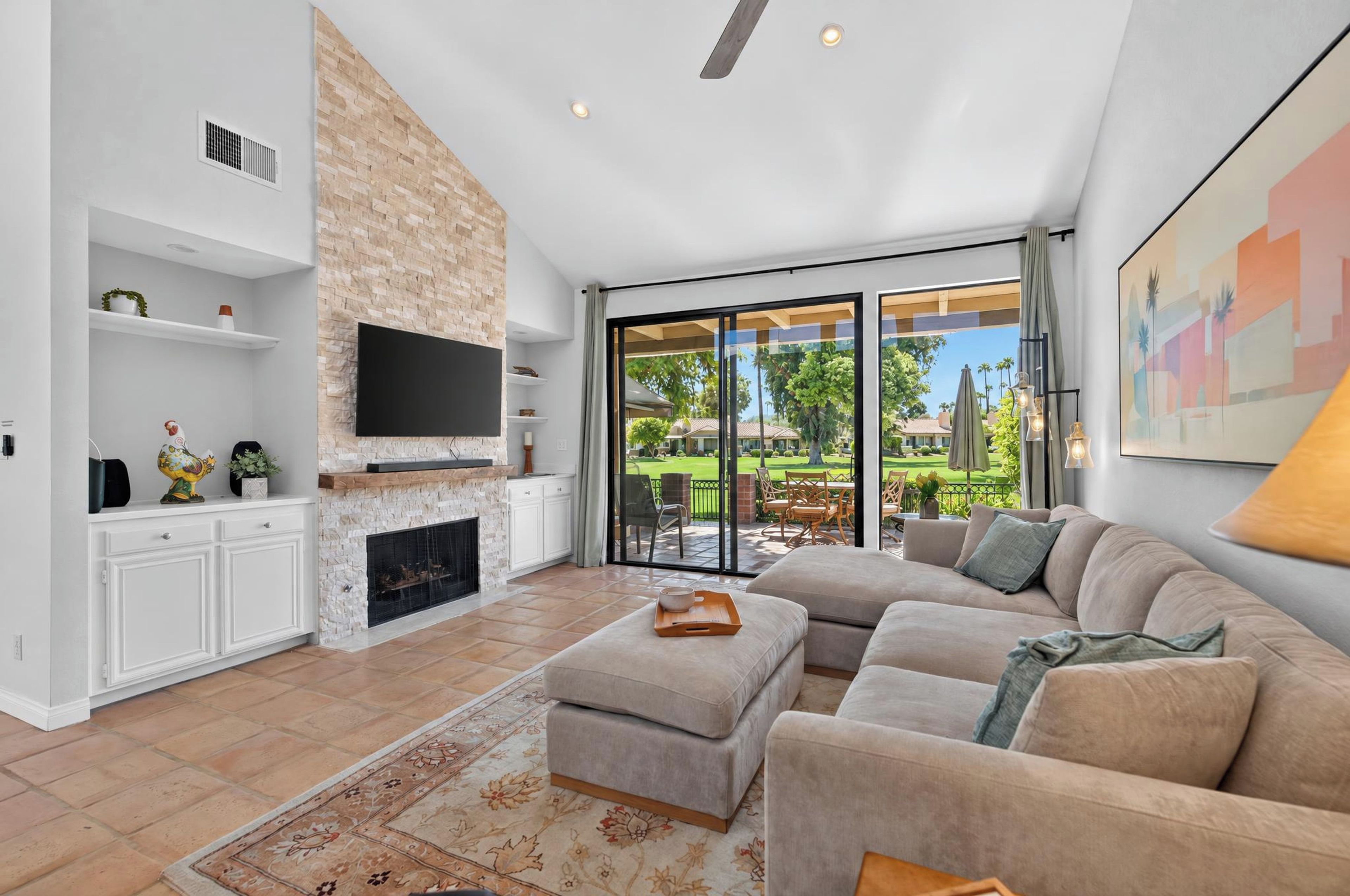 A spacious living room with a fireplace, a large flat-screen TV, a comfortable sofa, and a view of a well-maintained garden through large sliding glass doors.