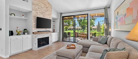A spacious living room with a fireplace, a large flat-screen TV, a comfortable sofa, and a view of a well-maintained garden through large sliding glass doors.
