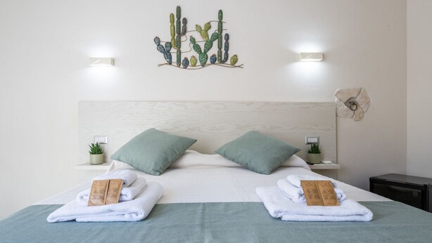 A neatly made bed with two pillows, a cactus wall art piece, and two bedside tables with small plants. - Il Ginepro - Suite Cactus (Pula)
