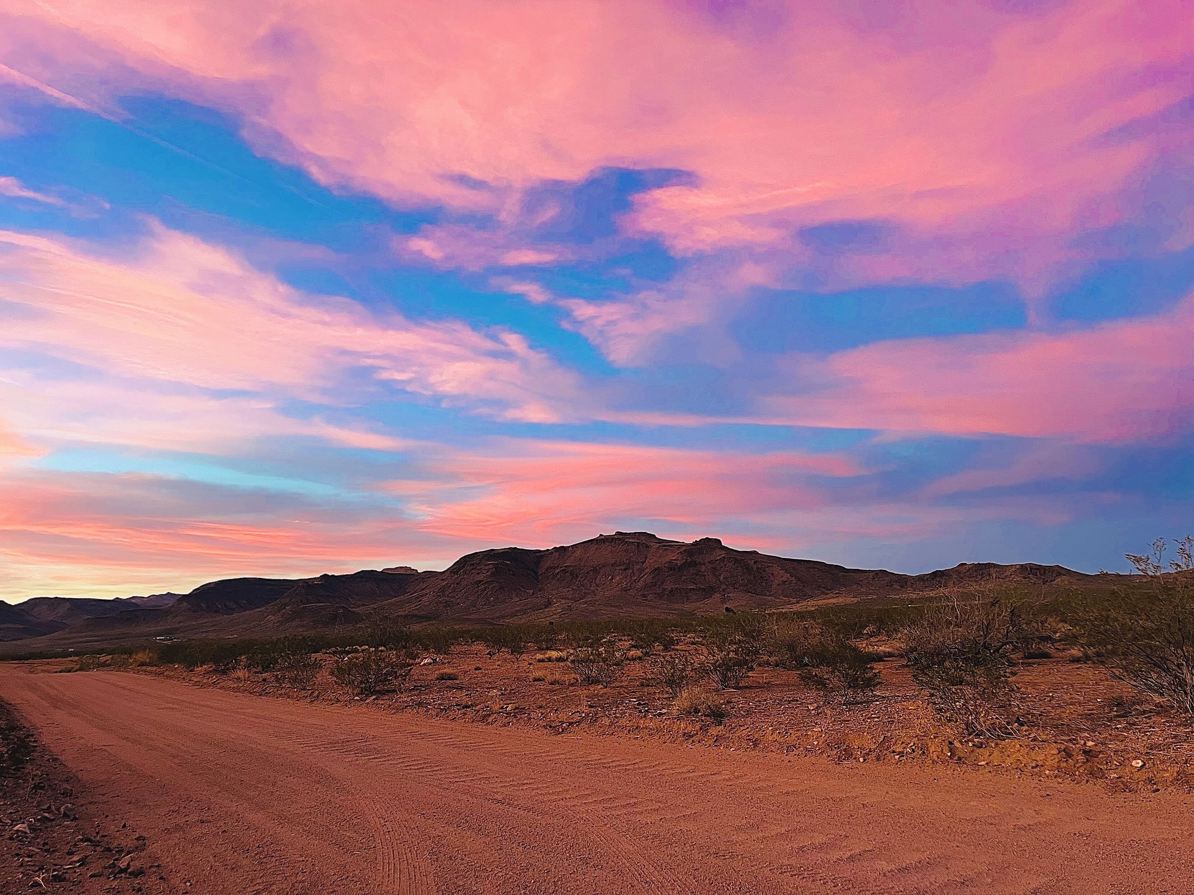 Paisaje desértico con un camino de tierra que se dirige hacia una cadena montañosa bajo un cielo colorido.